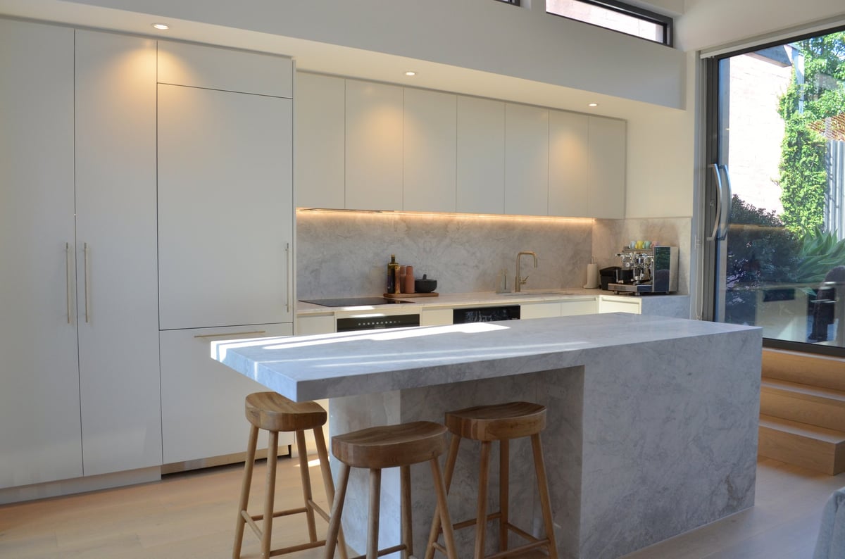 Modern minimalist kitchen with white cabinetry, concrete island counter, wooden bar stools, and floor-to-ceiling windows overlooking greenery