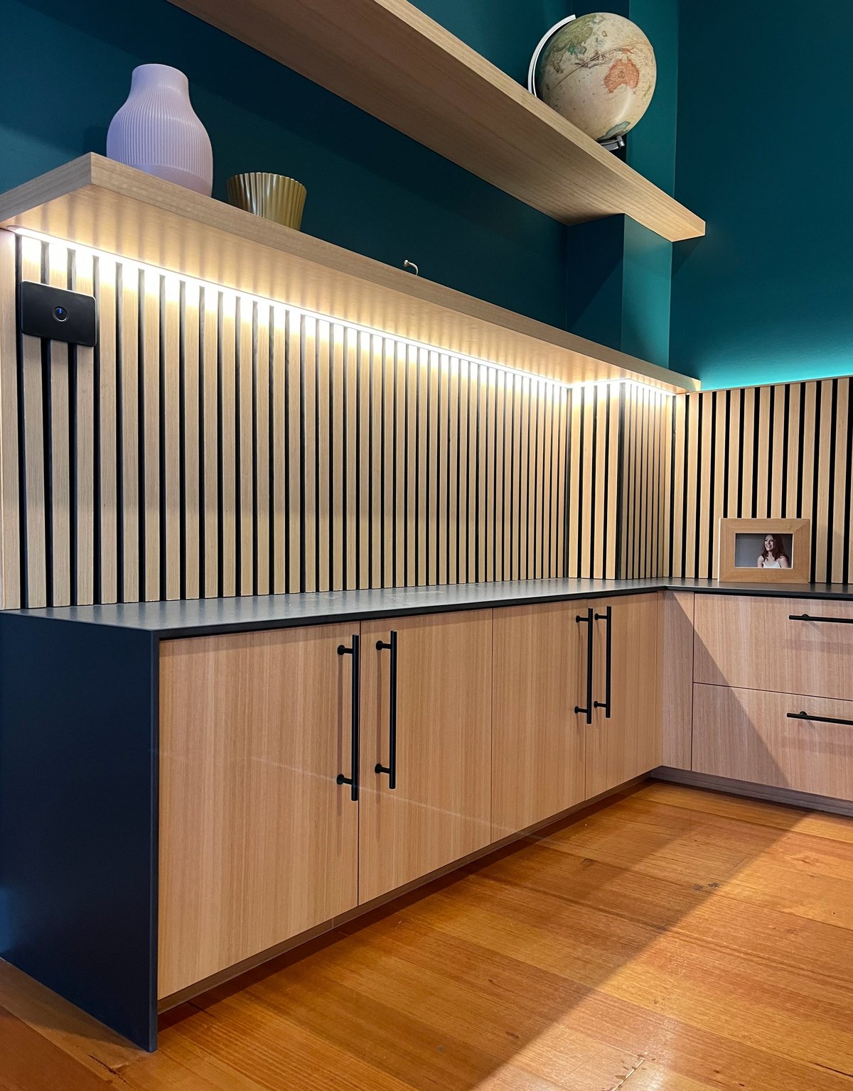 Modern kitchen with wood cabinetry, lit vertical slat backsplash, floating wooden shelf with decorative globe, and teal wall backdrop